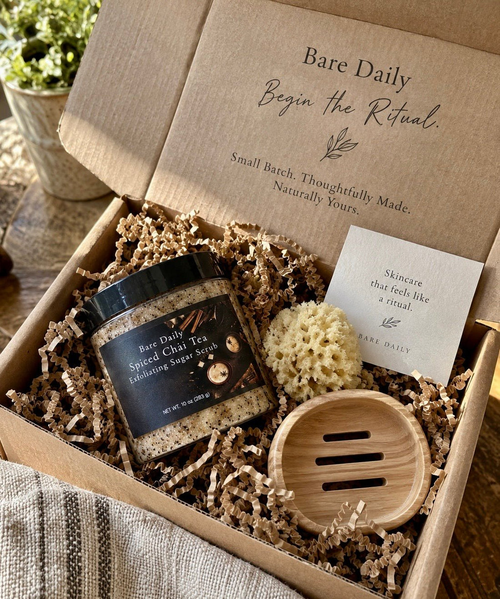 An open Bare Daily gift box filled with shredded paper, containing a jar of Spiced Chai Tea cleansing soap, a small beige scrubber, a wooden soap dish, and a branded card. The box lid shows the Bare Daily branding and slogan.