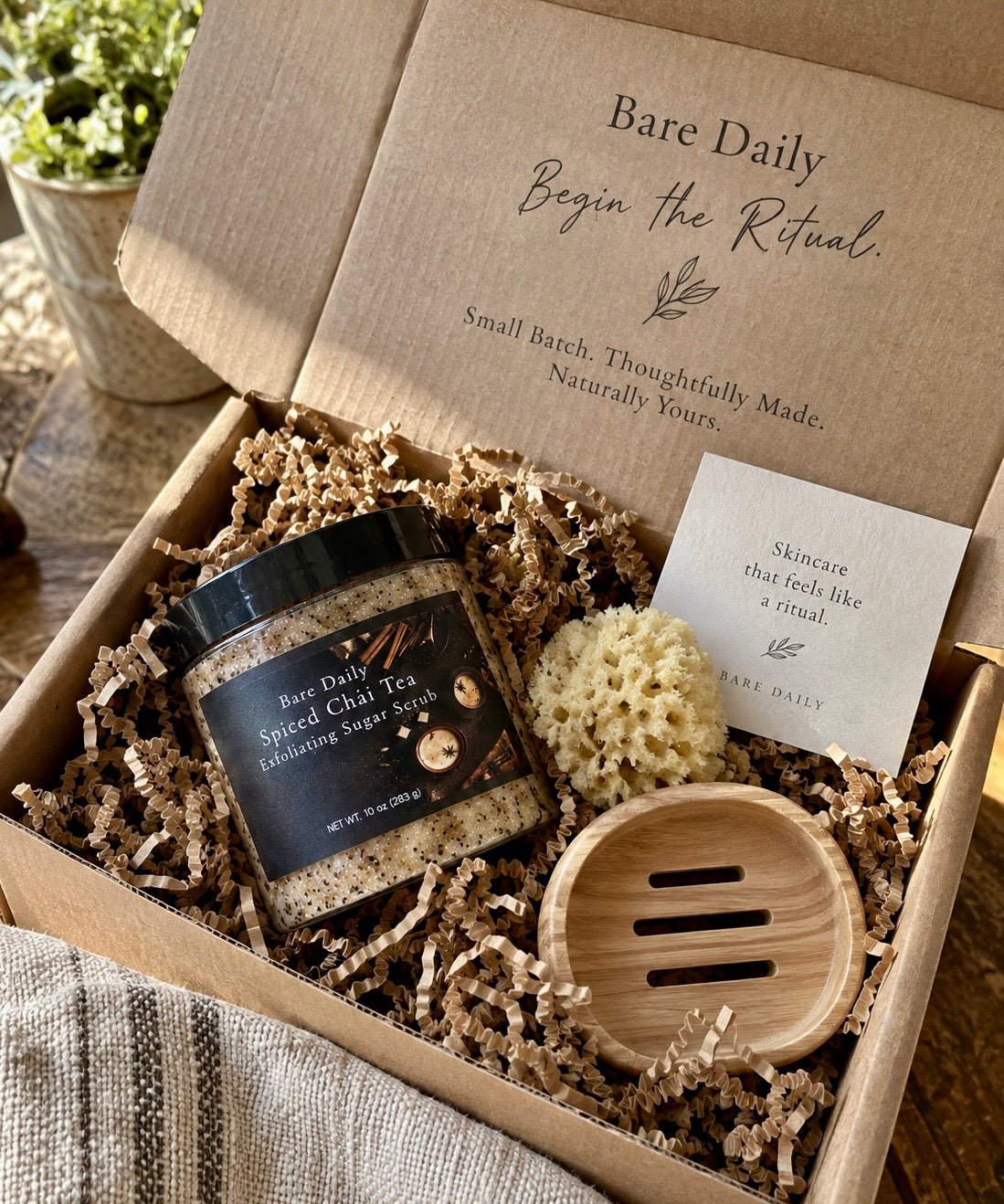 An open Bare Daily gift box filled with shredded paper, containing a jar of Spiced Chai Tea cleansing soap, a small beige scrubber, a wooden soap dish, and a branded card. The box lid shows the Bare Daily branding and slogan.