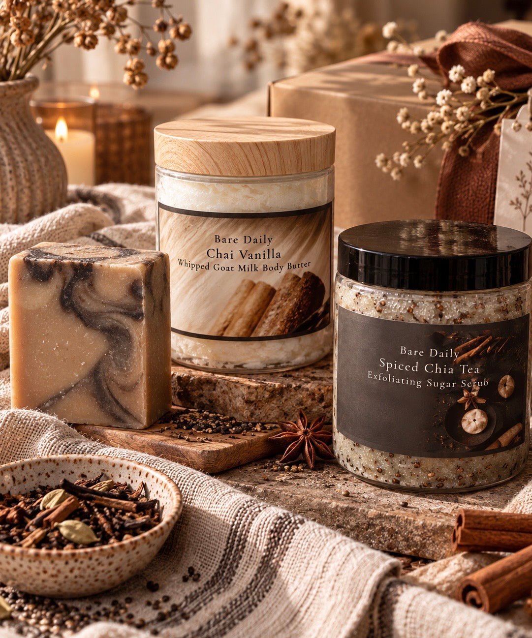A styled product display featuring a bar of marbled soap, a candle labeled Bare Daily Chai Vanilla, and a sugar scrub jar labeled Bare Daily Spiced Chia Tea, arranged on a rustic wooden surface with cinnamon sticks, a bowl of spices, and a wrapped gift box in the background.