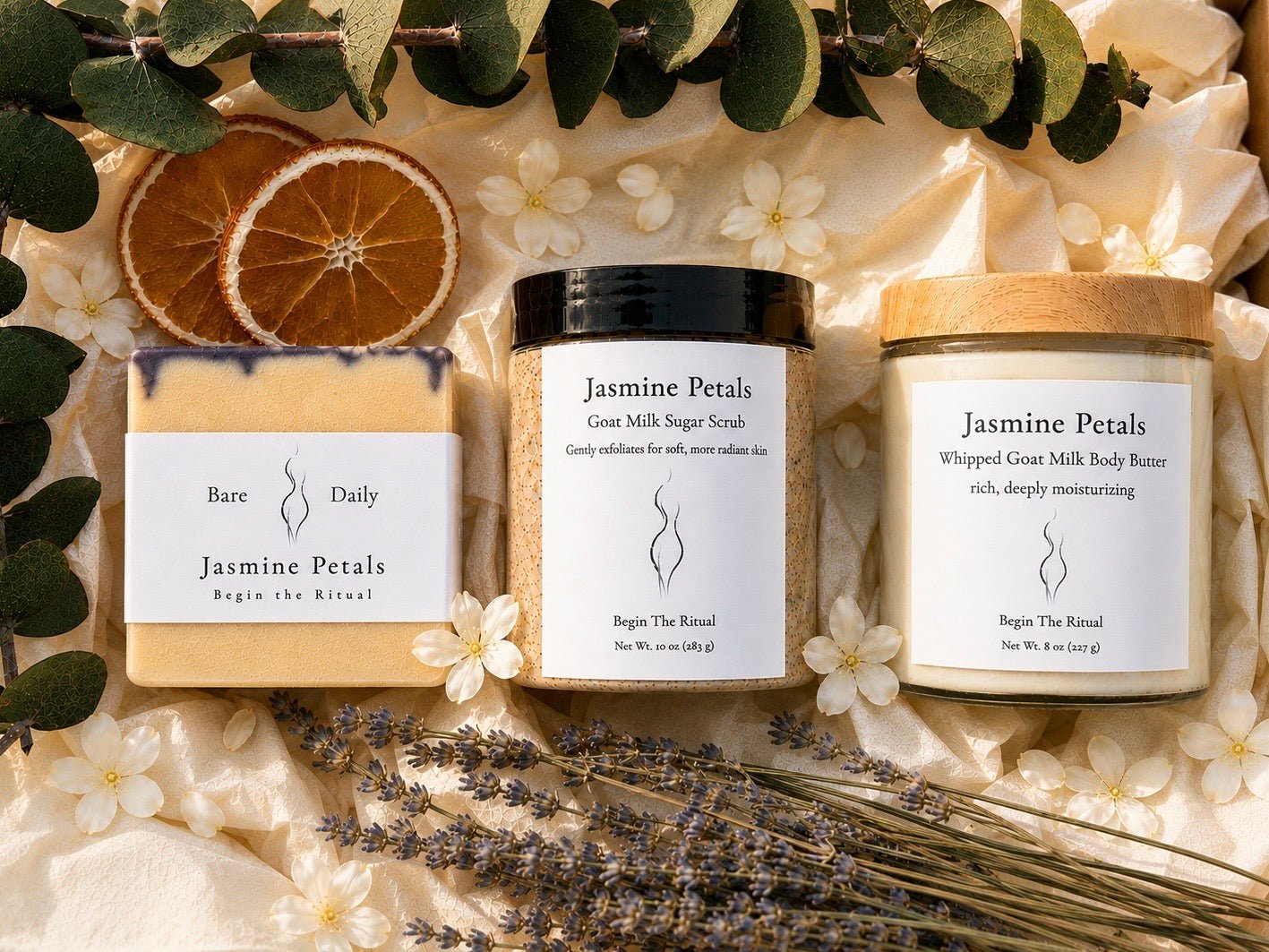 Three skincare products from Begin The Ritual are arranged in a styled flat lay with dried orange slices, white flowers, and lavender stems on cream fabric. The products include a bar of soap labeled 'Jasmine Petals Daily Jasmine Petal Bar', a jar of 'Jasmine Petals Great Milk Sugar Scrub', and a jar of 'Jasmine Petals Whipped Great Milk Body Butter'.