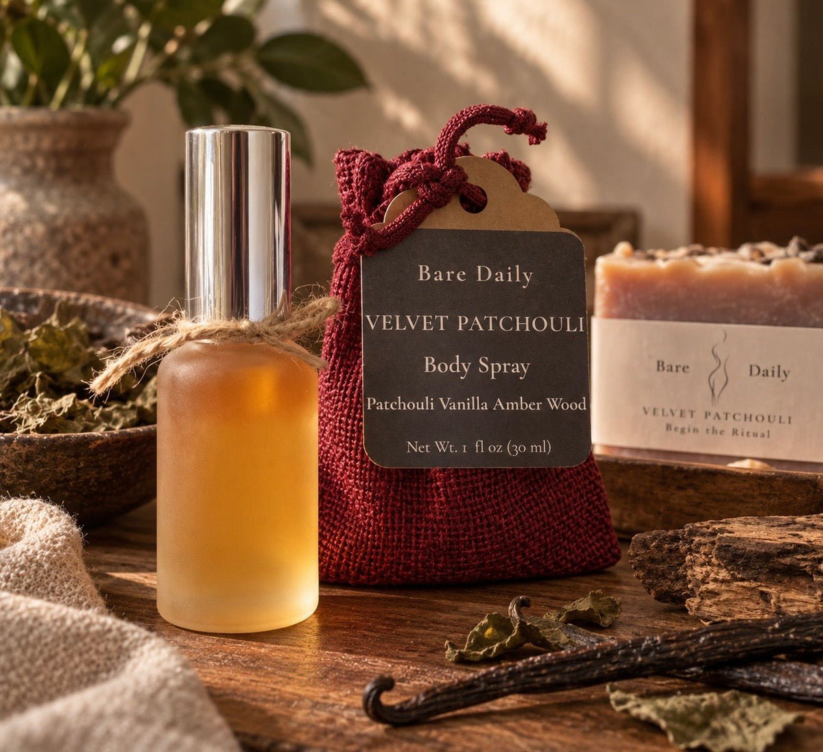A bottle of Bare Daily Velvet Patchouli Body Spray stands next to its dark red drawstring pouch packaging on a rustic wooden table, with dried herbs, a vanilla bean, and another soap bar in the background.