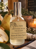 A frosted glass room and linen spray bottle with a silver cap, labeled 'Bare Daily FROSTED PEAR ORCHARD', styled with pears, a vanilla bean, and a lit candle in a cozy autumn setting.