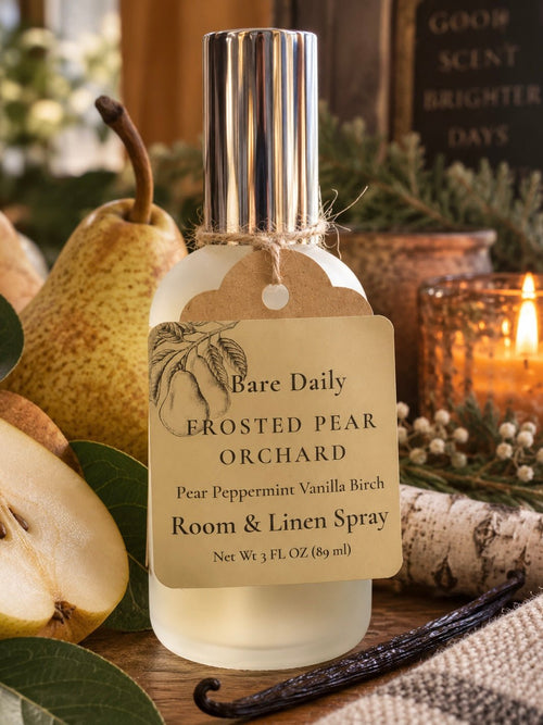 A frosted glass room and linen spray bottle with a silver cap, labeled 'Bare Daily FROSTED PEAR ORCHARD', styled with pears, a vanilla bean, and a lit candle in a cozy autumn setting.
