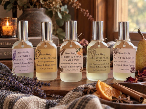Five Bare Daily room and linen spray bottles arranged on a wooden surface with seasonal botanical props including lavender, cinnamon, dried orange peel, and a candle in the background.