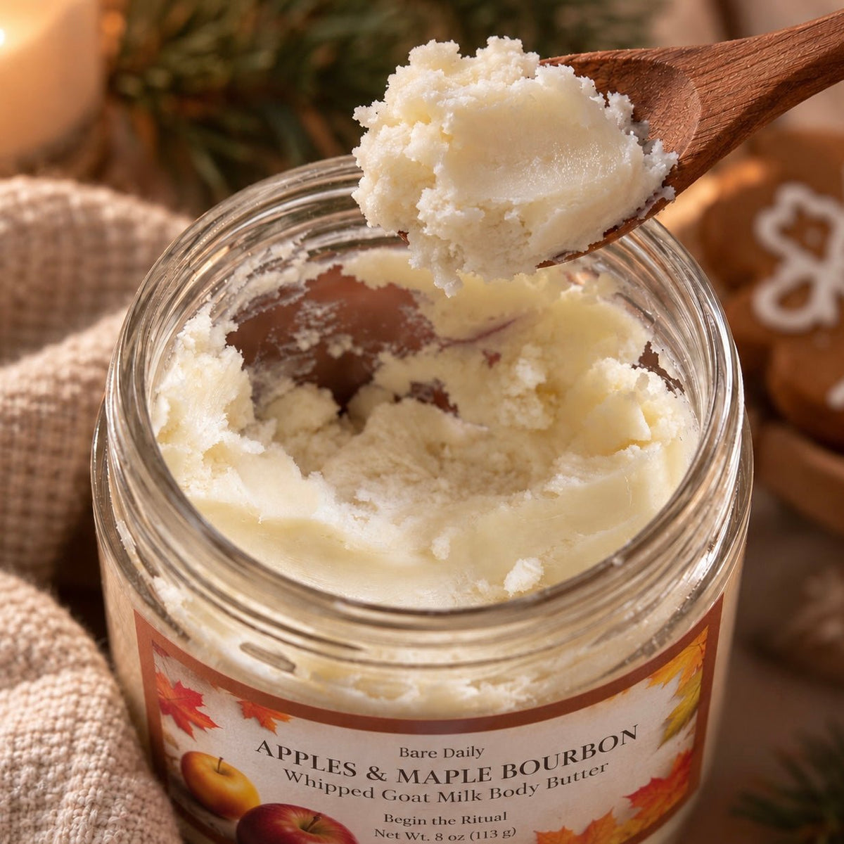 A glass jar of Bare Daily Apples & Maple Bourbon whipped goat milk body butter with a wooden spoon lifting cream-colored balm, set against a cozy autumn background with burlap, pine branches, and gingerbread cookies.