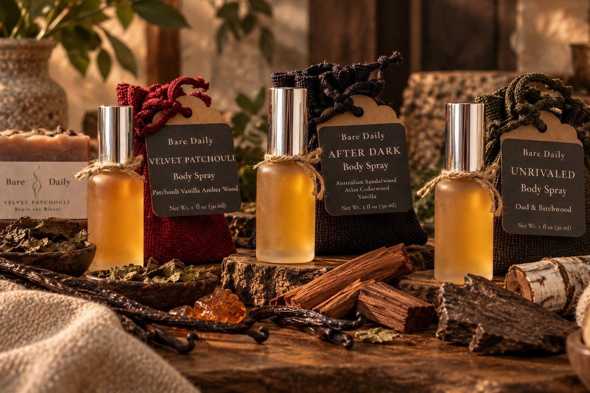 Four Bare Daily body spray bottles with silver caps and black labels are arranged on a rustic wooden surface with dried herbs, bark, and fabric. The amber-colored sprays come in scents including Velvet Patchouli, After Dark, and Unrivaled.