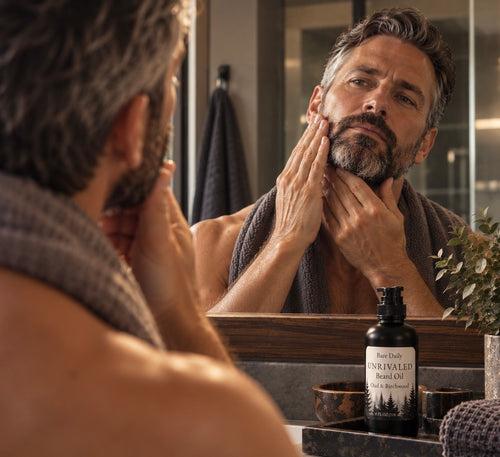 A well-groomed man applying Bare Daily Unrivaled Beard Oil to his beard and jawline in a bathroom mirror, showcasing a premium men's self-care and grooming routine.