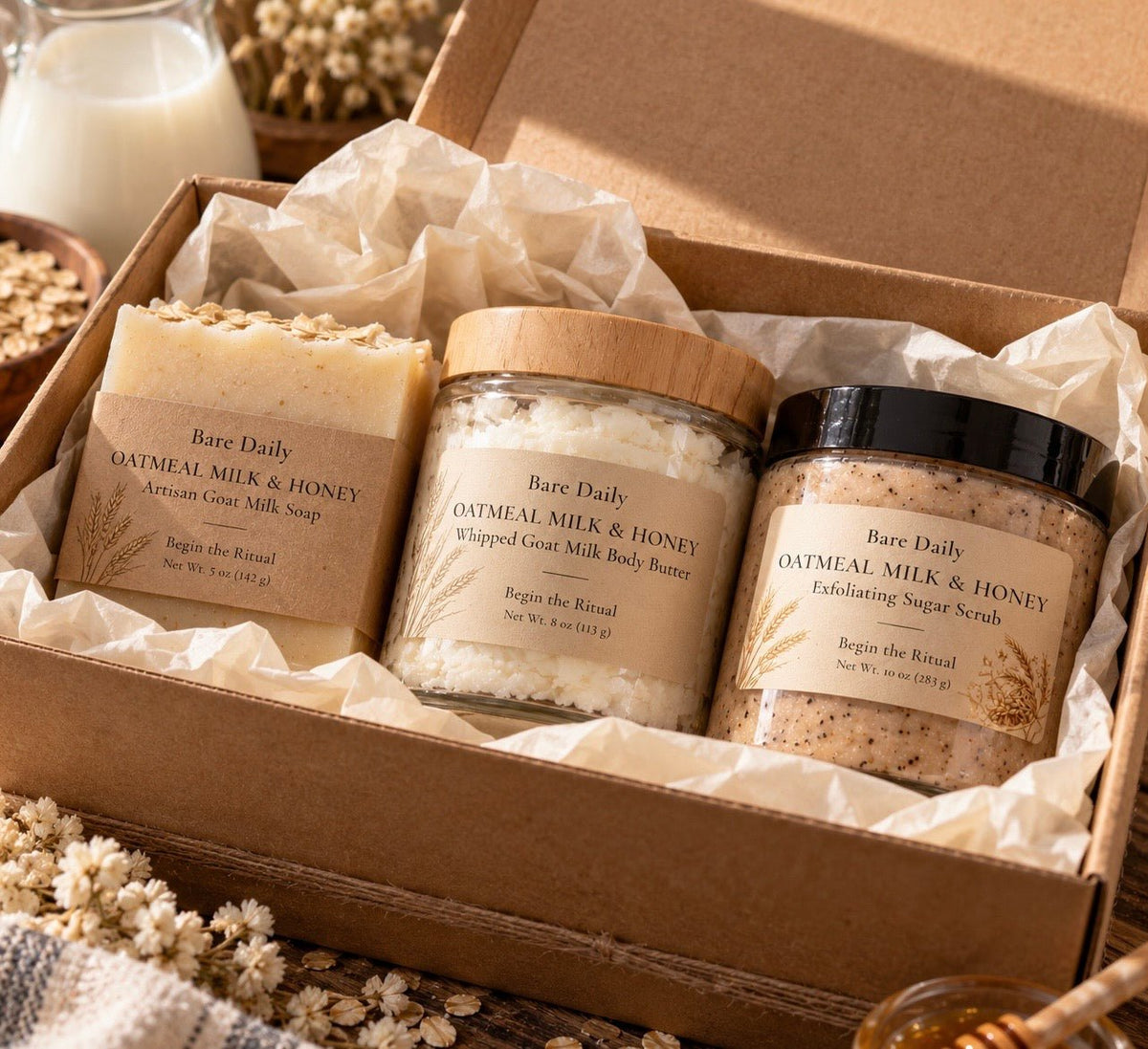 A gift box set containing three skincare products from Bare Daily: Oatmeal Milk & Honey Artisan Goat Milk Soap, Whipped Goat Milk Body Butter, and Exfoliating Sugar Scrubs, arranged in a kraft cardboard box with tissue paper.