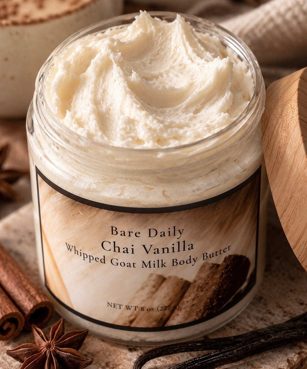 An open jar of Bare Daily Chai Vanilla Whipped Goat Milk Body Butter, showing the thick whipped cream product inside. The jar has a label with a warm wood-grain design, and it is styled on a rustic surface with cinnamon sticks, star anise, and a vanilla bean.