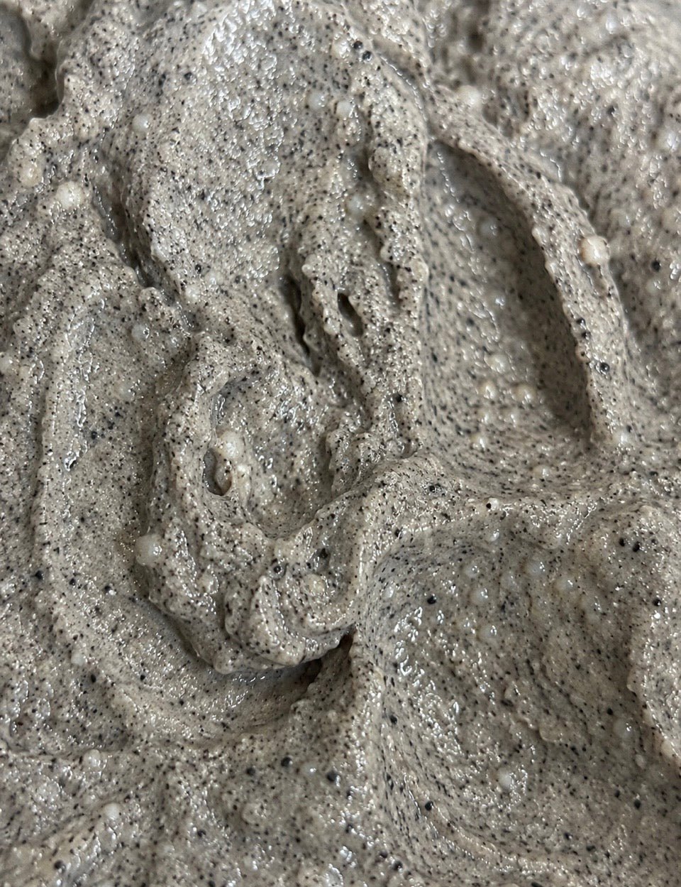 A close-up texture shot of a thick, swirled gray cosmetic cream or scrub with fine black and white specks, showing a rich, creamy consistency.