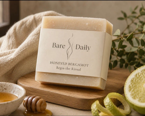  (labeled bar on wood board, honey dipper, sliced bergamot, eucalyptus, linen) Bare Daily Honeyed Bergamot Ancestral Lipid Bar with "Begin the Ritual" label on a wood cutting board, styled with a honey dipper, small bowl of raw honey, sliced bergamot, and eucalyptus sprigs on a linen cloth in warm natural light