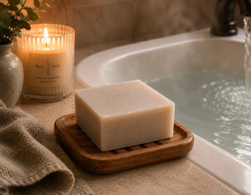 (unlabeled bar on wood soap dish beside filling bathtub, Bare Daily candle, linen towel, bronze faucet) Bare Daily Honeyed Bergamot Ancestral Lipid Bar on a ridged wood soap dish on a stone bathtub ledge beside a filling tub, with a lit Bare Daily branded candle, a linen towel, and a bronze faucet in warm ambient light