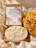 A bar of Bare Daily Rose & Honey goat milk facial cleansing bar displayed in front of its kraft paper packaging box, styled with an ethically sourced natural sea sponge and dried baby's breath flowers on a wooden self-draining natural Alder soap dish sourced in the Pacific NW.