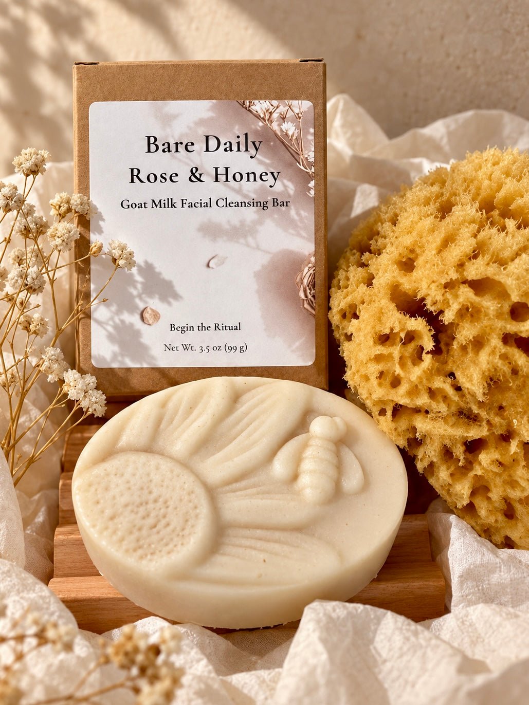 A bar of Bare Daily Rose & Honey goat milk facial cleansing bar displayed in front of its kraft paper packaging box, styled with an ethically sourced natural sea sponge and dried baby's breath flowers on a wooden self-draining natural Alder soap dish sourced in the Pacific NW.