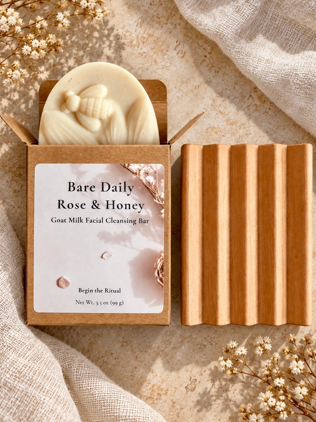 A boxed facial cleansing bar product labeled 'Bare Daily Rose & Honey' shown with the soap partially pulled from the kraft paper box and a self-draining Alder wood soap dish beside it on a textured fabric surface with dried baby's breath flowers.