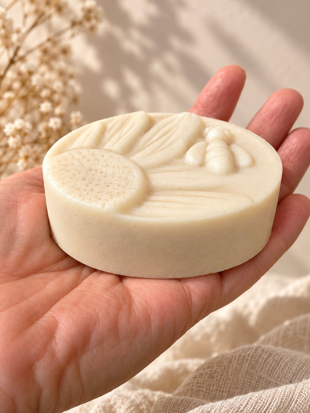 A hand holding a round, off-white syndet goat milk facial cleansing bar by Bare Daily with embossed floral and leaf patterns, set against a soft, warm neutral background with dried flowers.