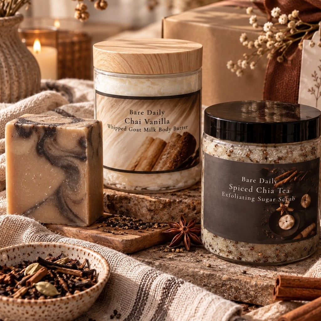 Two Bare Daily skincare jars and a handmade soap bar styled on a rustic wooden surface with spices, a candle, and wrapped gift boxes in the background. The products are Bare Daily Chai Vanilla Whipped Goat Milk Body Butter and Bare Daily Spiced Chia Tea Exfoliating Sugar Scrub.