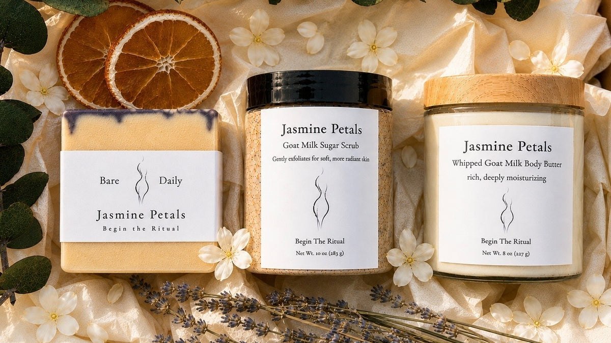 Three skincare products from Begin The Ritual are arranged in a styled flat-lay with dried orange slices, white flowers, and lavender sprigs. The products include a Bare Daily soap bar, a Jasmine Petals Goat Milk Sugar Scrub in a black-lidded jar, and a Jasmine Petals Whipped Goat Milk Body Butter in a wood-lidded jar.