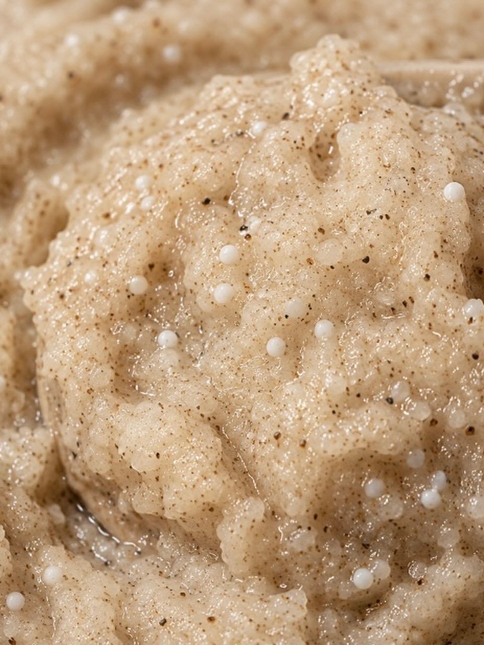 A close-up texture shot of a beige, creamy, grainy paste or scrub containing small white oval seeds and dark specks, filling the entire frame.
