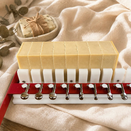 A set of handmade pale yellow bars of soap arranged in a white and red soap cutter or mold with metal guides, set on a beige fabric background with a small soap dish in the background.